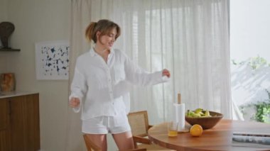 Dansçı kadın mutfak masasında taze portakal yiyor. Beyaz şortlu oyuncu kız ev mutfağında ritme göre hareket ediyor. Mutlu güzel bayan yalnız eğleniyor favori şarkının tadını çıkarıyor..