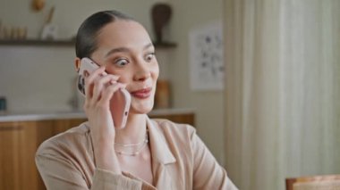 Neşeli bayan cep telefonunu arıyor modern mutfağı kapatıyor. Konuşan gülümseyen genç kadın şaşırmış görünüyor. Esmer kız evde akıllı telefondan iletişim kuruyor. Duygularını gösteriyor..