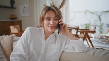 Gülümseyen kız rahat ev atmosferinde cep telefonuyla iletişim kuruyor. Mutlu kadın akıllı telefonu arıyor rahat koltukta tek başına oturuyor. Güzel, neşeli bir bayan cep telefonuyla konuşuyor, koltukta dinleniyor..