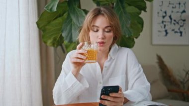 Kaygısız kadın meyve suyu içiyor. Ev masasında cep telefonu ekranına bakıyor. Beyaz gömlekli rahat bayan sabah kahvaltısında akıllı telefon çağrısına cevap veriyor. Güzel kız taze içecekten hoşlanıyor..