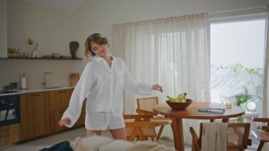 Sabah rutininde kulaklıkla dans eden mutlu kadın. Beyaz gömlekli ve şortlu genç bayan modern mutfakta kablosuz kulaklıkla müzik dinliyor. Hoş bir kız, evinde enerjik bir şarkı dinliyor..