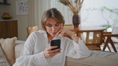 Cep telefonuna bakan düşünceli bir yüz ifadesiyle koltukta oturan odaklanmış bir kadın. Sakin bir aile sabahında akıllı telefon mesajı okuyan ciddi bir kadın. Çekici kız akıllı telefon ekranı.