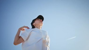 Meditasyon yapan, doğa üzerinde egzersiz yapan, kollarını esneten, şapka takan bir kadın. Motive olmuş kadın bulutsuz mavi gökyüzünün önünde spor yapıyor. Sıska sporcu kadın güneşli sokakta sağlıklı vücudu ısıtıyor..