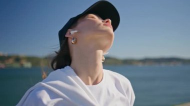 Güneşli nehir kıyısında boynunu uzatan kapalı gözlü sporcu kadın. Ciddi kadın sporcu yaz sabahı açık havada spor yapıyor. Aktif kız, kablosuz kulaklık takarak spor yapmayı seviyor..