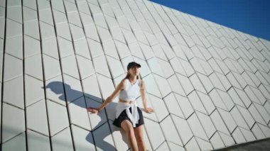 Sportif kadın, güneşli bir günde, beyaz geometrik duvarın yanında güven içinde poz veriyor. Rahat bir model modern mimariye dayanan kentsel yaz atmosferinin tadını çıkarıyor. Sıradan şehir ortamında rahatlayan şık bir sporcu.. 