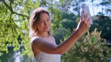 Neşeli kız güzel yeşillik doğasına telefon kamerası yerleştiriyor. Yaz gölü manzarasında akıllı telefon kullanarak fotoğraf çeken çekici bir kadın. Gülümseyen genç bir kadın güneş ormanında selfie çekiyor.