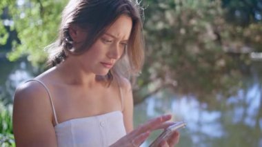 Güneşli bir parkta cep telefonu kullanan endişeli bir kadın. Tatminsiz genç bayan yeşil doğada akıllı telefon yazan bozuk aletle karıştırıyor. Sinirli kız cep telefonundan kötü haber alıyor.