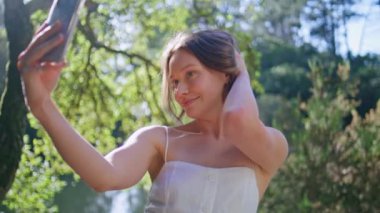 Flört eden kız, gölün kıyısında, yakın plan beyaz elbiseli bir şekilde poz vererek selfie çekiyor. Şirin, kibar, akıllı telefon kameralı bir kadın güneşli yeşilliğin önünde fotoğraf çekiyor. Şefkatli bayan içeriği kaydediyor 