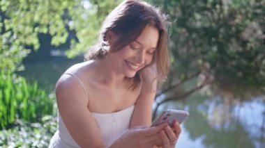 Güneşli parkta cep telefonuna göz atan neşeli bir kadın. Portre mutlu esmer, akıllı telefona odaklanmış ormandaki ağaçların keyfini çıkarıyordu. Huzurlu atmosferde cep telefonunu izleyen güzel kız.