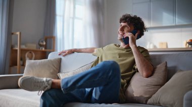 Konforlu oturma odasındaki kanepede akıllı telefonla iletişim kuran mutlu hipster. Gülümseyen adam hafta sonu cep telefonu paylaşımından bahsediyor. Dinlenen adam kanepede dinleniyor ve içeride keyifli sohbetler ediyor.