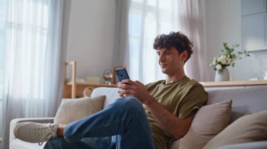 Evdeki adam cep telefonu uygulamasını inceliyor. Modern apartmandaki akıllı telefon ekranında oturan yakışıklı genç adam. Gülümseyen müşteri cep telefonu uygulaması kullanarak online sipariş veriyor 