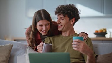 Modern dizüstü bilgisayarları kullanarak evde film izlerken gülen çiftler. Gülümseyen genç eşler kanepede rahat bir şekilde oturup kahve fincanı içerek komik filmlerin tadını çıkarıyorlar. Rahatlamış aile hafta sonu. Evlilik rutini 