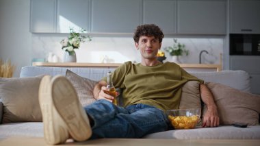 Relaxed man watching show on tv resting at home weekend. Smiling chilling guy drinking beer enjoying television movie on couch. Happy handsome hipster relaxing eating snacks at sofa. Homie lifestyle clipart