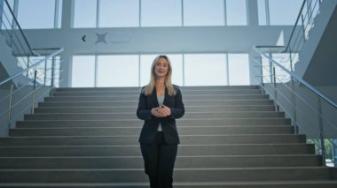 Businesswoman standing with outstretched arms on staircase corporate office. Smiling woman executive displaying confident posture on stairs. Ambitious stylish lady welcoming embodying leadership. clipart