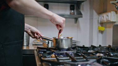 Modern mutfak fırınında sosu hazırlayan profesyonel bir aşçı. Bilinmeyen aşçı iş yerinde kırmızı domates sosunu tencereyle karıştırıyor. Mutfakta et yemeği için malzeme yapan restoran işçisi.
