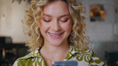Modern kafeteryada cep telefonu mesajı okuyan sevimli bir kadın. Kafe masasında oturan akıllı telefon görünümlü gülümseyen kadının portresi. Cep telefonunda sosyal medya içeriğine göz atan mutlu kız.