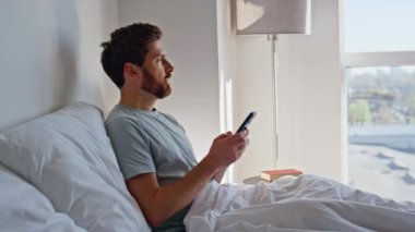 Uyanmış adam sabahın erken saatlerinde yatakta akıllı telefon kaydırıyor. Rahat bir adam, rahat yatak odasındaki beyaz yatağa uzanmış cep telefonuyla sosyal medyada geziniyor. Tembel evde hafta sonu cep telefonu kullanan sakin bir esmer.