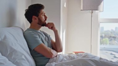 Hüzünlü adam, modern otel odasında yatarken mutsuz oluyor. Üzgün adam rahat yatak odasında rahat rahat düşünceli bir şekilde dinleniyor. Erkek esmer, sabah güneşinde beyaz battaniye içinde dalgın bir ifade takınıyor. 