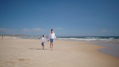 Kumsalda koşan neşeli anne çocuğu. Neşeli bir aile, güneşli okyanus dalgalarının tadını çıkarıyor. Mutlu anne erkek elini tutuyor ve yaz tatilini deniz kıyısında geçiriyor. Çocukluk anneliği kavramı.
