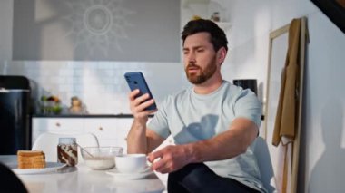 Sabahçı adam cep telefonuna mesaj atıp kahve fincanıyla tek başına oturuyor. Rahatlamış adam mutfakta akıllı telefondan mesaj okuyor gibi görünüyor. Neşeli serbest çalışan kapalı mekanda sosyal medya cep telefonunu kaydırıyor 