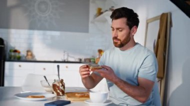 Kahvaltıcı adam açık mutfakta sandviç hazırlıyor. Sakallı, aç, lezzetli şeyler yiyen, sabah rutinini yalnız yapan adam. Tatmin olmuş esmer, en sevdiği yemeğin tadını çıkarıyor.