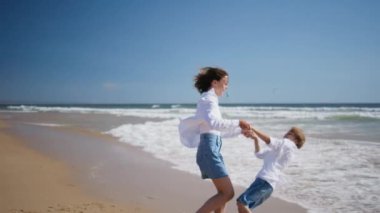 Anne köpüklü okyanus dalgalarına basan mutlu çocuğu döndürüyor. Güzel kumsalda eğlenen eğlenceli aile bağları. Gülümseyen anne, sevgili oğluyla yaz sahilinde enerjik bir aktivitenin tadını çıkarıyor..