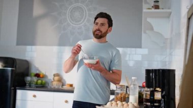 Kahvaltıda ayakta kahvaltı eden tatmin olmuş bir adam. Tıraşsız, gülen bir adam granola yiyor ve modern ışık evine bakıyor. Sakin Freelancer öğle yemeğini rahat bir evde tek başına yiyor. 