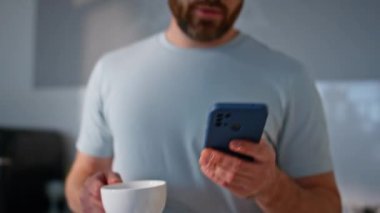 Açık mutfakta elinde akıllı telefondan kahve fincanı tutan adam elleri. Mutlu iş adamı mesajları okuyor ve espresso içiyor. Gülümseyen sakallı adam kahve yudumluyor lüks bir dairede cep telefonu izliyor.