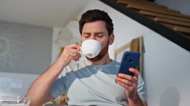 Kahvaltılık adam cep telefonunu kaydırıyor ve ev mutfağında kahve içiyor. Akıllı telefon mesajı okuyan mutlu adam yalnız başına vakit geçiriyor. Modern mutfakta rahatlamış erkek aromatik içeceği yudumluyor.