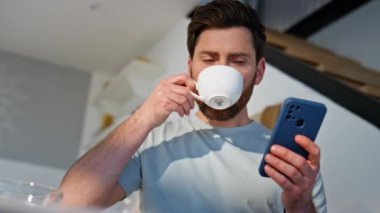 Sabahçı adam akıllı telefonları karıştırıyor. Mutfağı kapatıyorlar. Yakışıklı, aromatik kahve içen cep telefonu görünümlü bir adam. Dairede cep telefonu uygulaması kullanarak rahatlamış erkek alışverişi