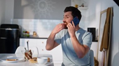 Kahvaltılık adam, akıllı telefondan arayıp mutfakta sandviç yiyor. Evde oturan rahat adam cep telefonuyla sabah keyfine bakıyor. Sakin erkek rahat evde telefonda haberleri tartışıyor.