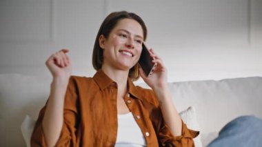 Gülümseyen ev hanımı cep telefonu dedikodusu yapıyor rahat, modern kanepeye yakın oturuyor. Neşeli bir kadın rahat koltukta rahat rahat cep telefonuyla konuşuyor. Kahkaha atan esmer daire odasında akıllı telefondan arıyor..