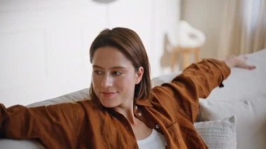 Kanepede oturan kadın beyaz yumuşak yastıkların keyfini çıkarıyor. Rahat bir kanepede yalnız başına rahatlayan gülümseyen çekici bir kadının portresi. Modern apartman dairesinde dinlenen mutlu esmer ev kadını..