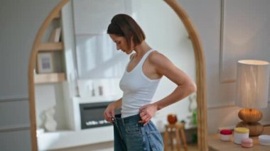 Çağdaş iç mekandaki yansımayı incelemeye çalışan üzgün kadın. Zayıf kadın, kilo vermiş, aynaya bakıyor. Mutsuz esmer, bol kot pantolon giyiyor ve canı sıkkın. Diyet sonucu