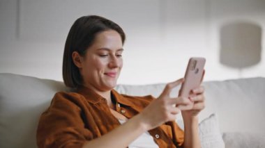 Kanepede rahat rahat oturup cep telefonuna mesaj atan ev kadını. Rahat ev atmosferinde rahatlarken akıllı telefonla mesajlaşan kaygısız gülümseyen bayan. İnternette telefonla sohbet eden mutlu kız öğrenci..