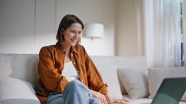 Videodaki neşeli kadın rahat koltukta oturuyor. Dizüstü bilgisayara el sallayan gülümseyen kadın kanepede online görüşmeye başlıyor. Dairedeki sanal konferansta pozitif kız konuşuyor..