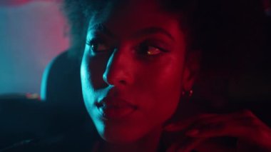 Portre kıvırcık saçlı model. Koyu renk araba pozu veriyor. Şehir ışıklarını yakıyor. Kırmızı neonun altında parlayan güzel Afro-Amerikan kadın. Ciddi kadın otomobil yolcusu tek başına pencereye bakıyor..