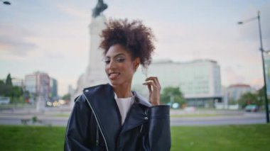 Kıvırcık kadın kamerada gülümsüyor bulutlu şehir caddesinde dikiliyor. Buklelerle oynayan mutlu Afrikalı Amerikalı kız portresi. Kaygısız moda kadını şehirde yalnız poz veriyor. Deri ceket giyiyor..