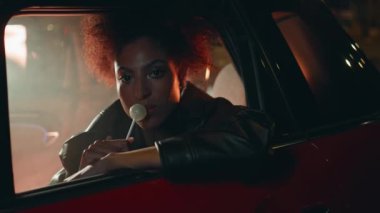 Parlayan gece aydınlatması altında araba camına yaslanmış şeker yiyen kendine güvenen bir kadın. Rahat Afro-Amerikan bayan otomobilin içinde lolipop yalıyor. Şehvetli esmer otomobilde şekerlerin tadını çıkarıyor..