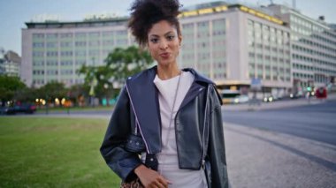 Pozitif esmer gezgin şehir hafta sonu alacakaranlık yakın çekim keyfini çıkarıyor. Gülümseyen Afro-Amerikan kadın modern caddede yürüyen kameraya bakıyor. Çekici kıvırcık kadın geceyi şehir meydanında yalnız geçiriyor..