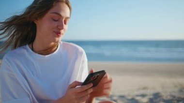 Plajdaki kadın, Oceanside 'da akıllı telefonlara mesaj atıyor. Çekici bayan güneşli bir hafta sonunda cep telefonu kullanarak kumlu kıyıda dinleniyor. Güzel rüzgarlı saç modeli internette geziniyor. Tatil yaşam tarzı kavramı 