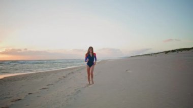 Günbatımı plajında koşan kaygısız sporcu kadın. Mutlu seksi kadın, kumsalda ayak izleri bırakarak huzurlu kıyı şeridinden zevk alıyor. Sakin deniz dalgalarında dinlenen neşeli sörfçü kadın neşe dolu bir akşam. İyi hissediyorum. 