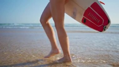 Sörfçü kızın bacakları okyanus kıyısında su basıyor. Okyanusa giren tanınmayan sporcu kadın aktif hafta sonunun tadını çıkarıyor. Yalın ayaklı kadın yaz sahillerine gidip tropik ada tatiline çıkıyor.