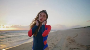 Kumsalda yürüyen sporcu kadının sabah portresi. Koruyucu takım elbiseli sevimli genç bir kadın. Kameraya bakıyor. Güneşin doğuşunu seyrediyor. Rüzgarlı, güzel kız kumlu kıyıya çıkıyor. Huzurlu doğada yalnız poz veriyor. 