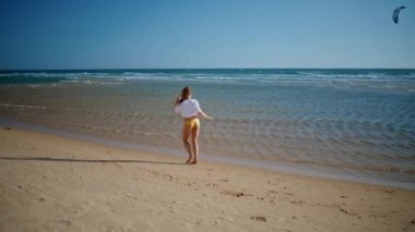 Sahilin arka manzarasında koşan kaygısız bir kız. Mutlu seksi kadın sarı bikinili huzurlu kumsal şeridinde eğleniyor. Sakin deniz dalgalarında dinlenen neşeli turist bayanın tatili sevindirici. İyi hissediyorum. 