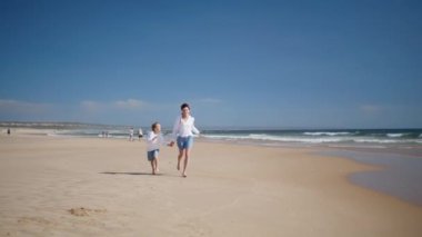 Kumsalda koşan neşeli anne çocuğu. Neşeli bir aile, güneşli okyanus dalgalarının tadını çıkarıyor. Mutlu anne erkek elini tutuyor ve yaz tatilini deniz kıyısında geçiriyor. Çocukluk anneliği kavramı.