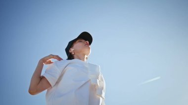 Meditasyon yapan, doğa üzerinde egzersiz yapan, kollarını esneten, şapka takan bir kadın. Motive olmuş kadın bulutsuz mavi gökyüzünün önünde spor yapıyor. Sıska sporcu kadın güneşli sokakta sağlıklı vücudu ısıtıyor..