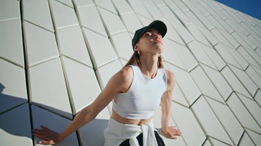 Duvara yaslanan atletik kadın modern yapı sakinliğini yakın çekim hissediyor. Şapka takan şık bayan yaz güneşinin tadını çıkarıyor. Kentsel geometrik mimaride fitness odaklı genç model.