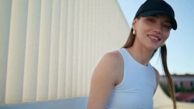Şehir korkuluklarında poz veren neşeli bir kadın neşeli bir gülümsemeyle kameraya bakıyor. Siyah şapkalı çekici esmer tek başına şehirde dikiliyor. Mutlu bayan dışarıda idman yaptıktan sonra dinleniyor..
