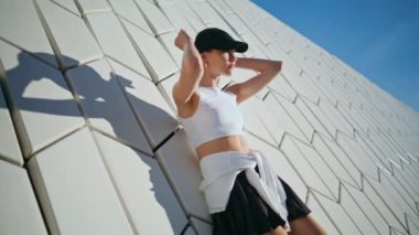 Duvarda poz veren spor modelinin geometrik dokusu güneş ışığının altında güneşleniyor. Modern şehir mimarisine dayanan güzel bir atletik kadın. Siyah şapkalı şık bayan, ellerini kaldırarak güneşli günün tadını çıkar..